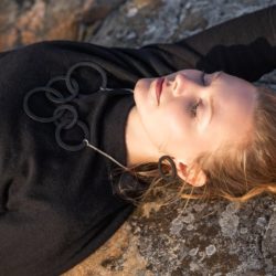 Nainen makaa silmät kiinni kalliolla päällään musta paita ja puinen kaulakoru ja korvakorut