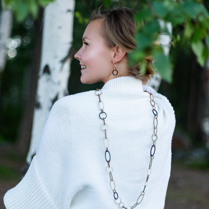 Vaalea nainen koivikossa valkoisessa neuleessa puinen ketjukaulakoru selässä