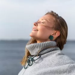 Nainen silmät kiinni aurinkoon päin, korvissa pyöreät vihreät korvakorut, kaulassa rengasmainen kaulakoru, harmaa villapaita